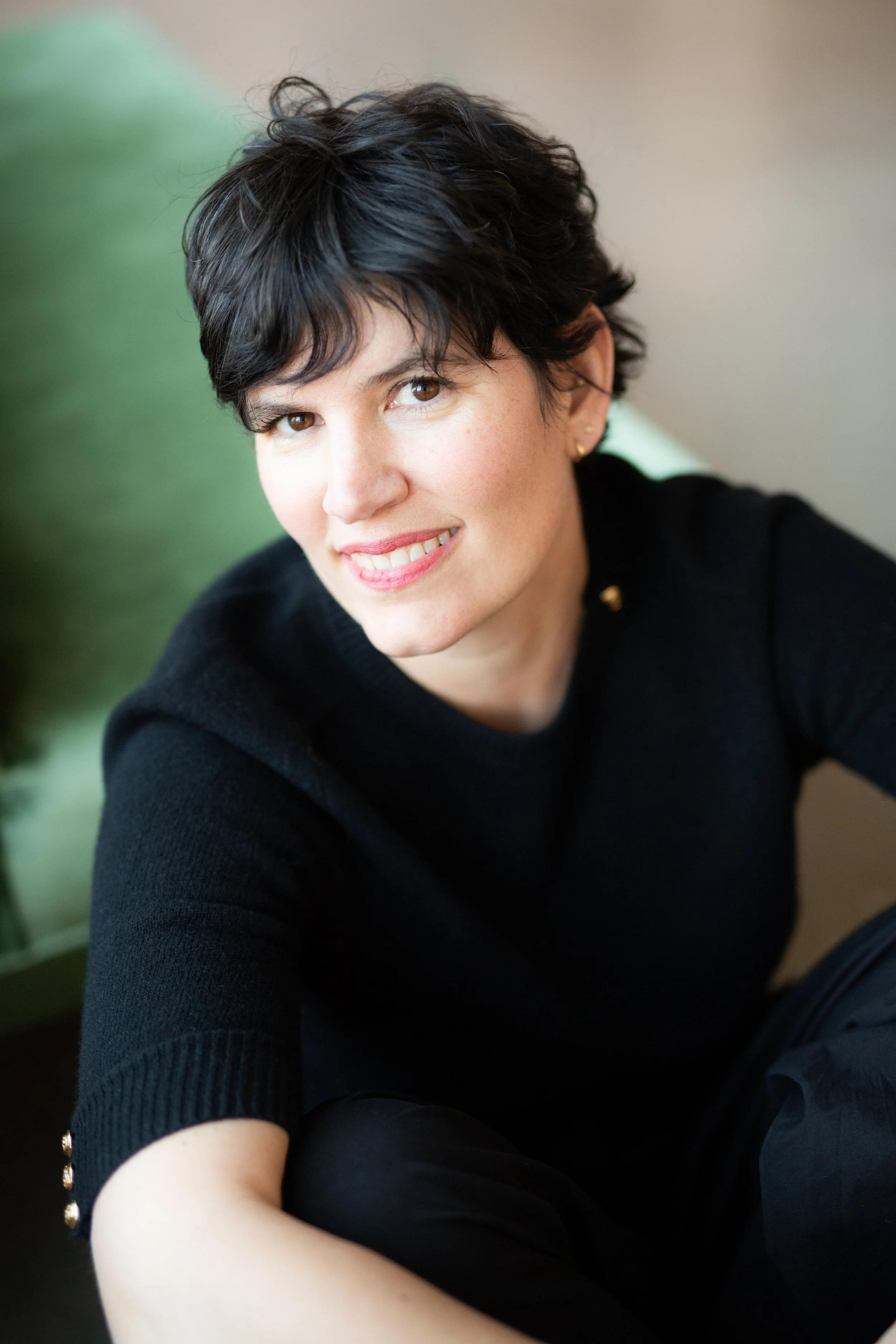 A light skinned woman sitting in front of a green chair. She has short black hair and is wearing an elbow-length black shirt with gold buttons on the sleeves.