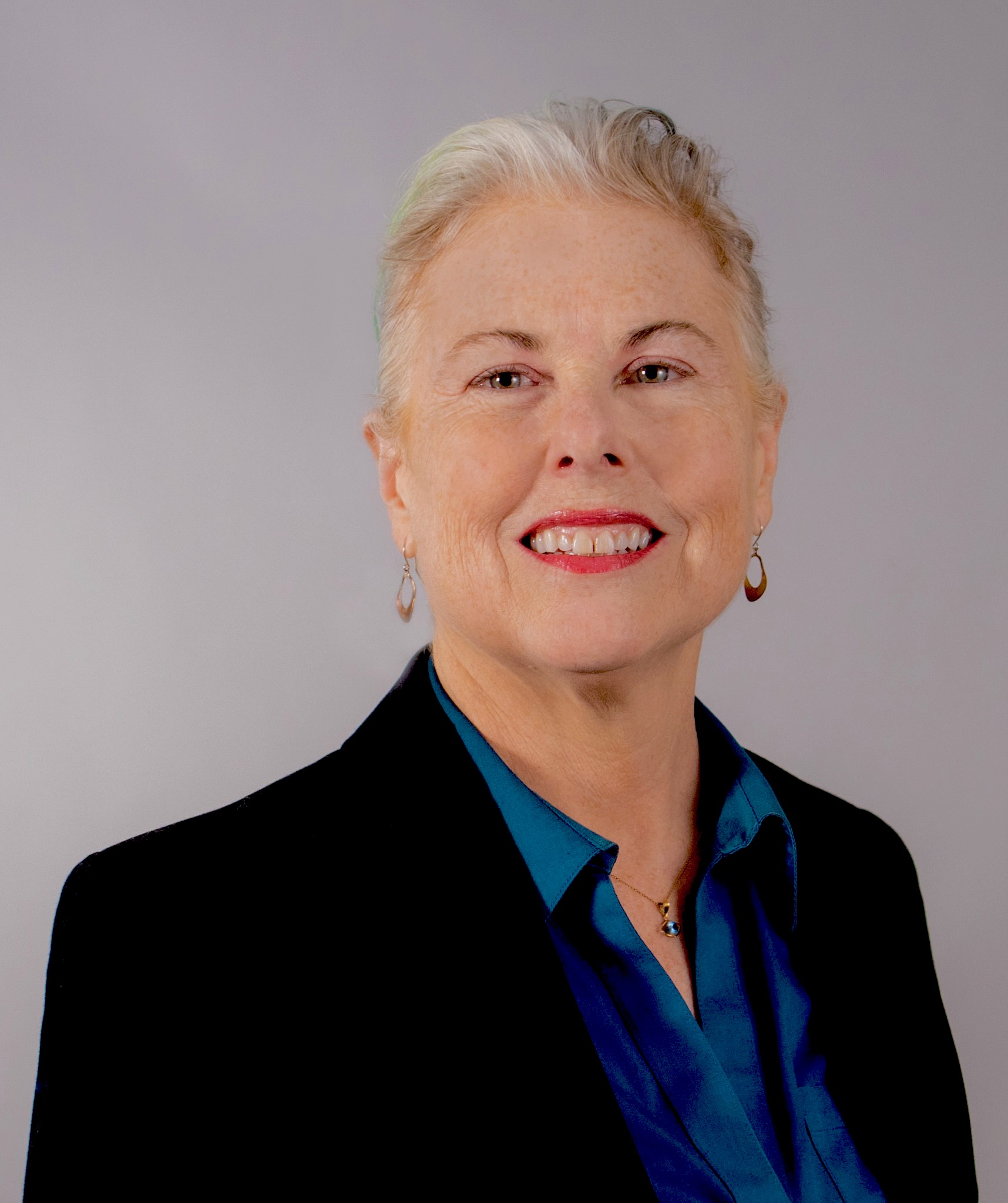 : A light-skinned woman photographed from the chest up in front of a white background. She has short white-blonde hair and is wearing a black blazer over a blue button-down shirt.