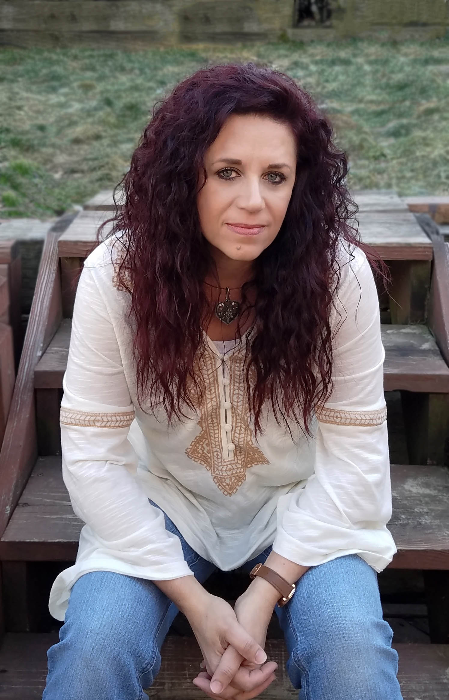 Liz is sitting on wooden steps. Liz has long, dark purple-reddish wavy hair and is wearing a long sleeve tunic, blue jeans, a heart shaped necklace, and a watch.