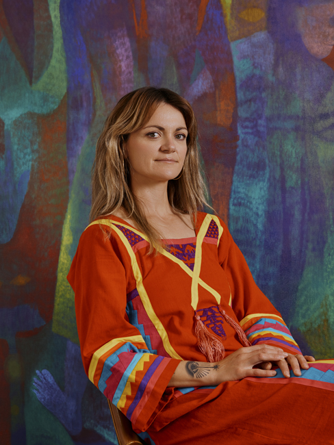A light-skinned woman sits in front of a cool-toned abstract background. She has blonde hair past her shoulders, and she is wearing a red dress multicolored sleeves and yellow fabric intersecting over the chest.