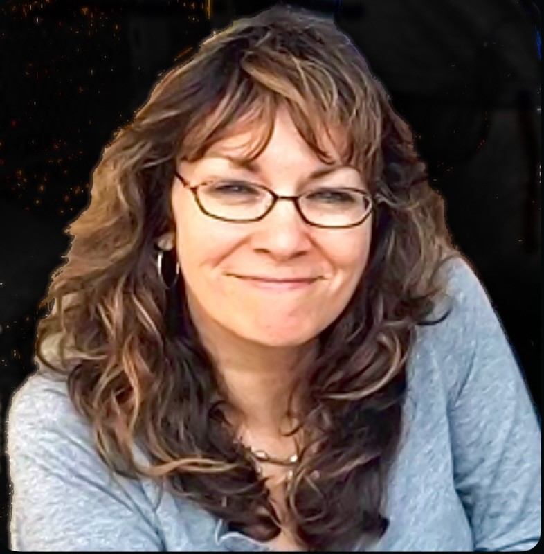 Mari is seen with long brown wavy hair and glasses. Mari is wearing a light blue shirt and has a gentle smile on her face.