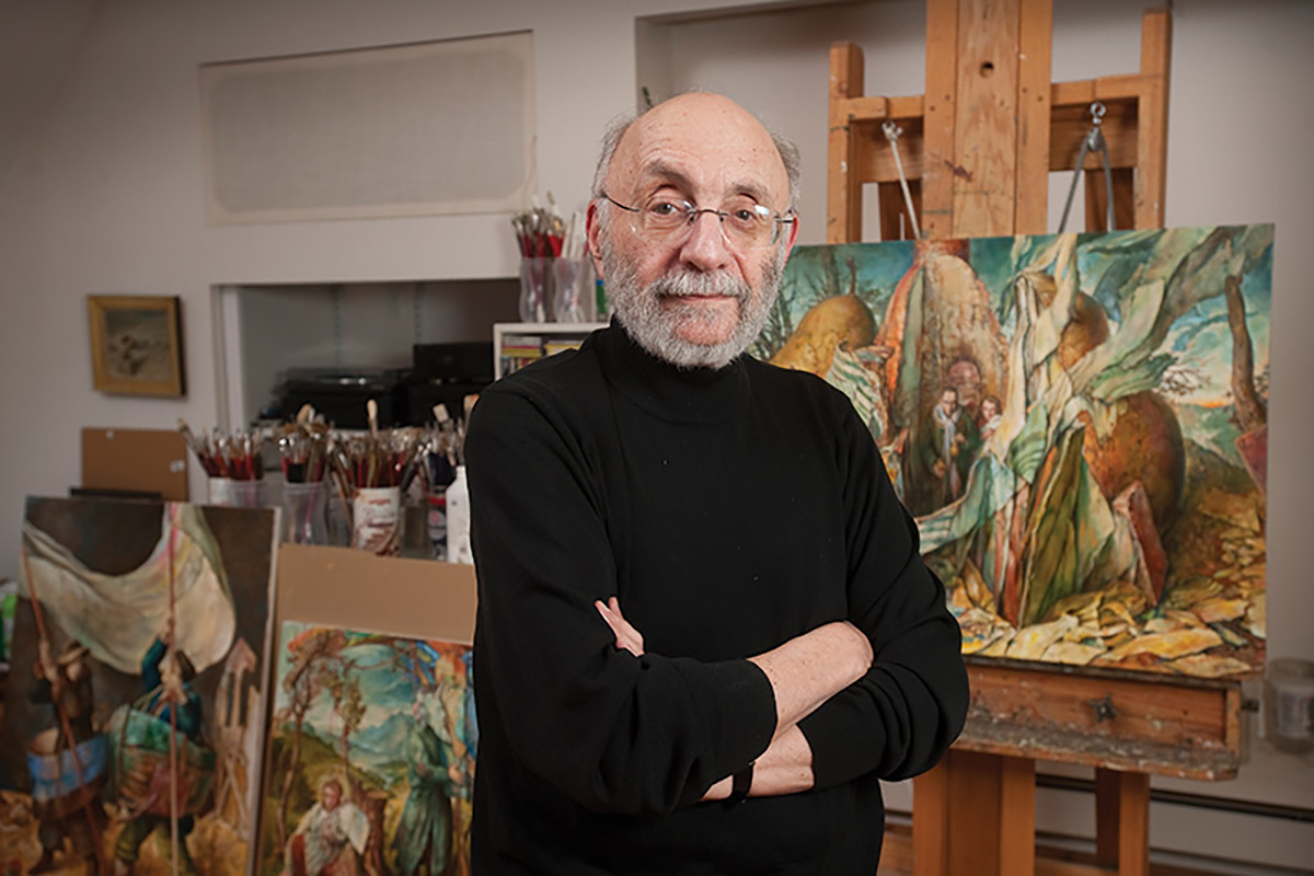 A light-skinned older man standing in an art studio while crossing his arms. He has grey hair on the side of his head, a grey beard, and rimless glasses. He is wearing a black turtleneck.