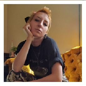 Viy is seen sitting on a vintage sofa with their right arm on their knee propping their head up, right hand pressed against their cheek. Viy has short blonde looking hair, facial piercings, and is wearing a tee-shirt with pants that have a light and dark pattern . 