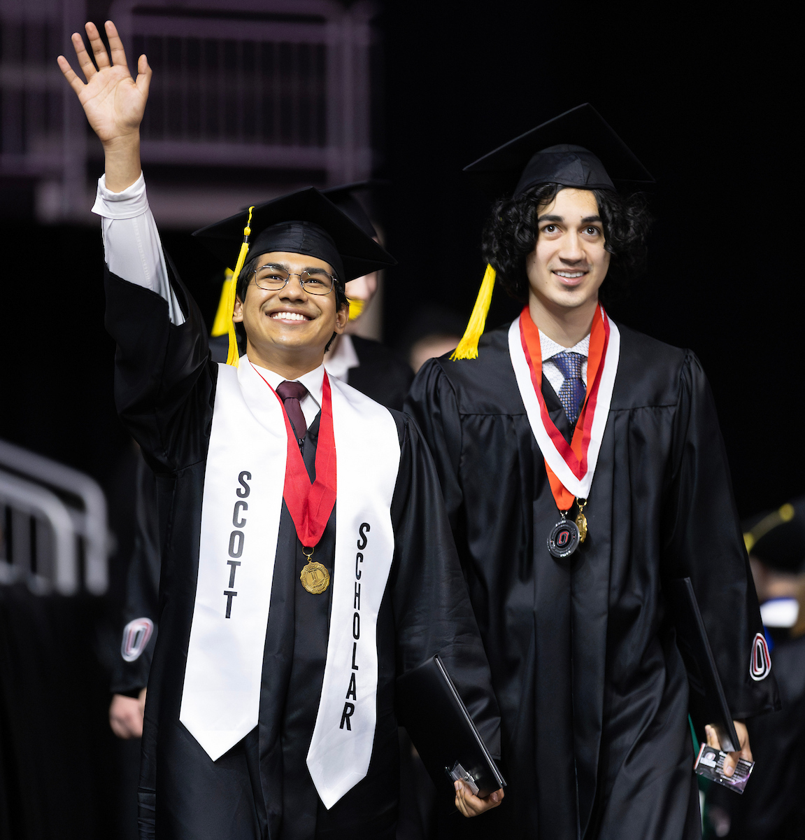 two male graduates, one wearing a scott scholars stole and waving to the crowd walk through baxter arena