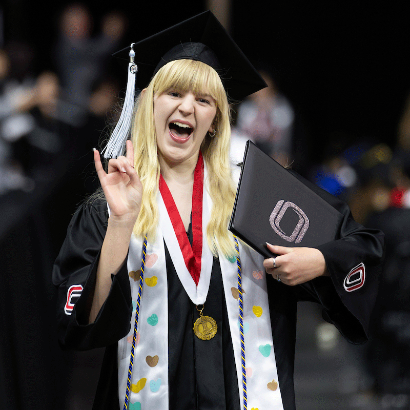 a woman with long yellow hair smiles and holds her diploma while flashing maverick horns