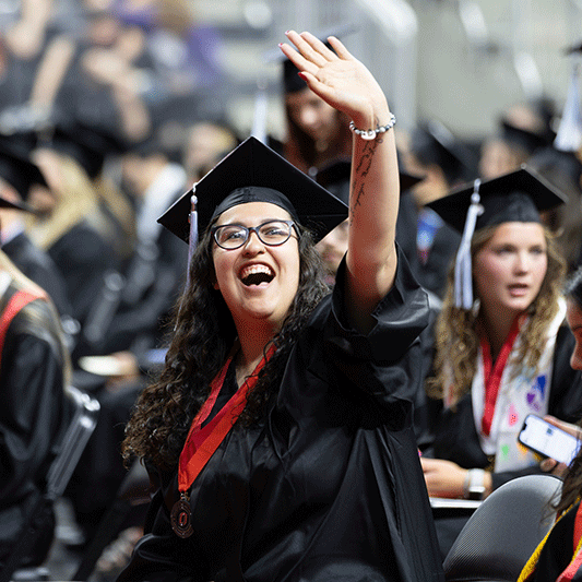 a woman with dark curly hair and glasses smiles and waves to her people in the crowd at uno's commencement