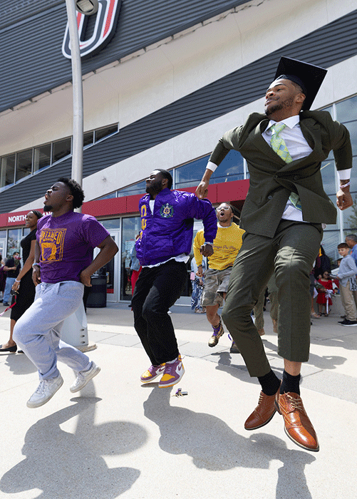 a group of black men in fraternity colors and a commencement gown jump and dance outside of baxter arena