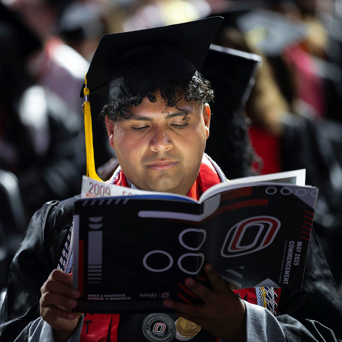 a young man of color dressed in his graduation gown sits and reads uno's commencement program