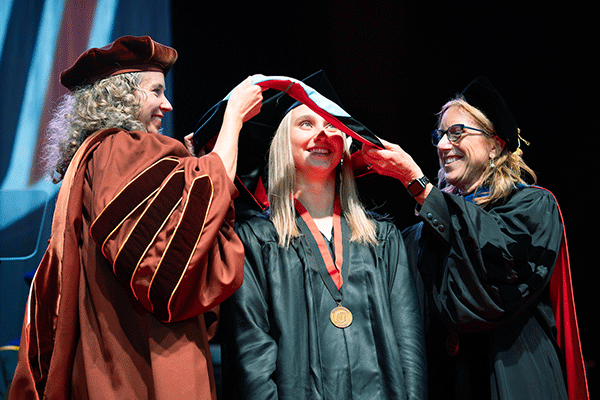 a woman with blond hair smiles as she is hooded while receiving her master's degree on stage at uno
