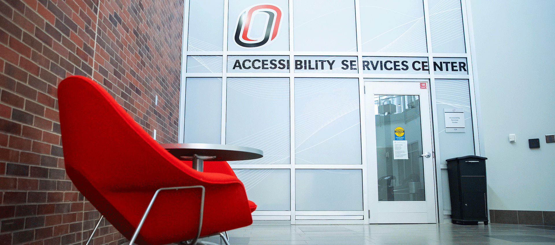 Three university students and staff stand close together, smiling inside a decorative red, black, and gold balloon arch while another person takes their picture at a campus celebration, with event photos visible in the background.