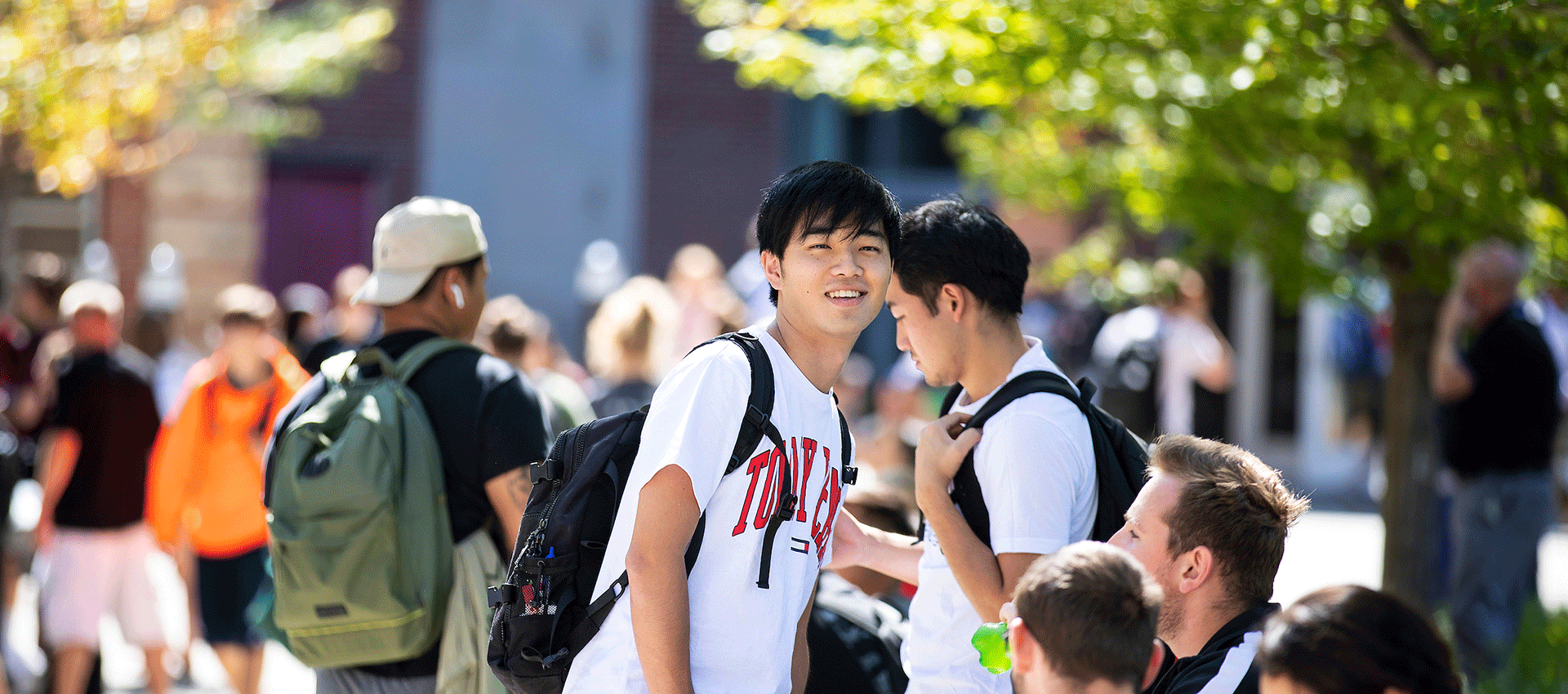 Students walk and gather along a tree-lined campus walkway between classes, with one student smiling toward the camera.