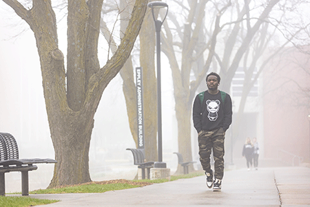 student walking on a college campus on a foggy morning, there are leafless trees and benches, the sidewalk looks wet