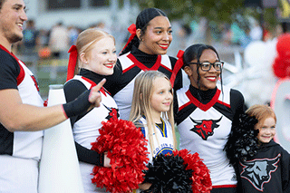 Members of a university cheer team in black, white, and red uniforms pose smiling with two children, holding pom-poms at a campus spirit event.