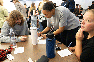 Students sit around a table while a staff member leans in to assist one student with paperwork, as others review forms and materials during an academic support event.