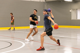 Students participate in a dodgeball game inside a campus recreation gym, with one player lunging forward to throw a ball while others prepare to dodge or catch.