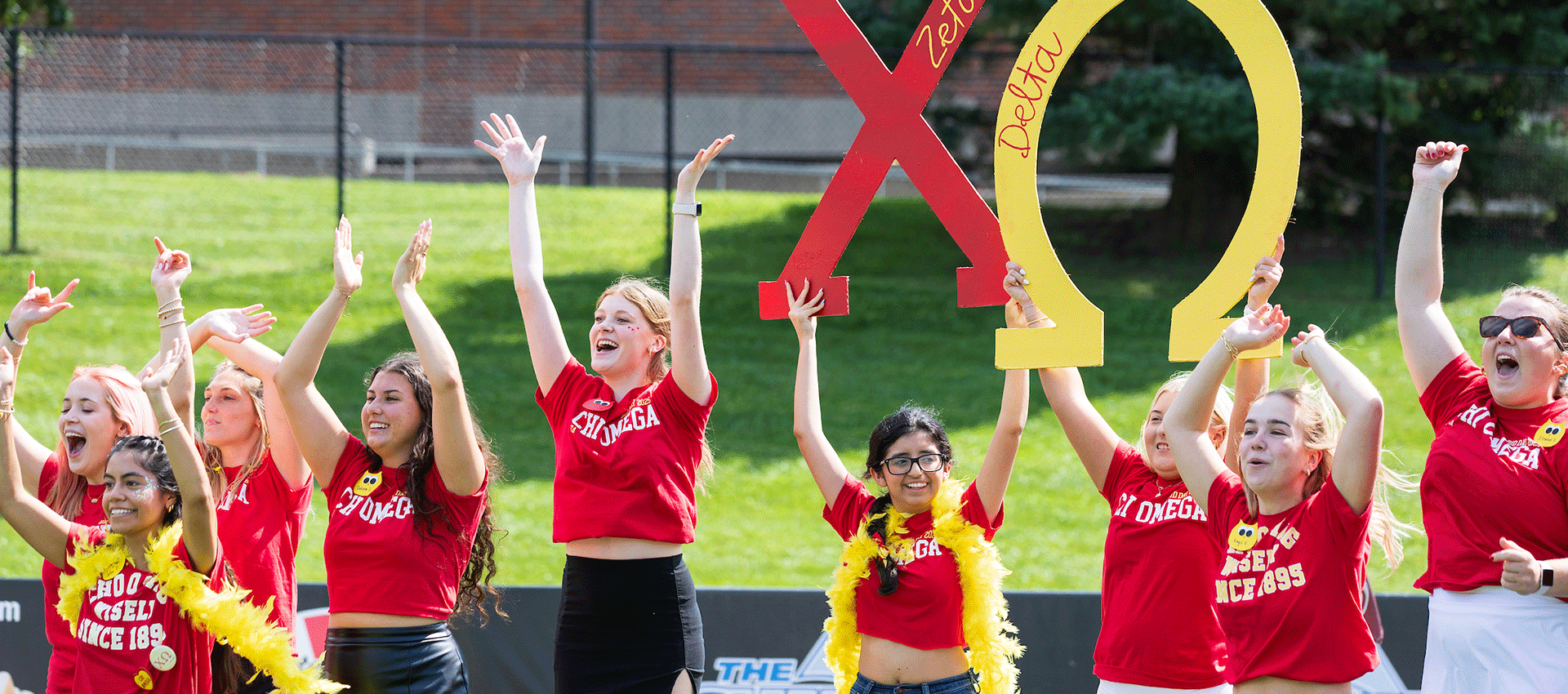 Three university students and staff stand close together, smiling inside a decorative red, black, and gold balloon arch while another person takes their picture at a campus celebration, with event photos visible in the background.