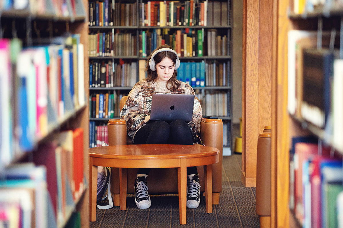 A student sits in a library chair wearing headphones while working on a laptop.