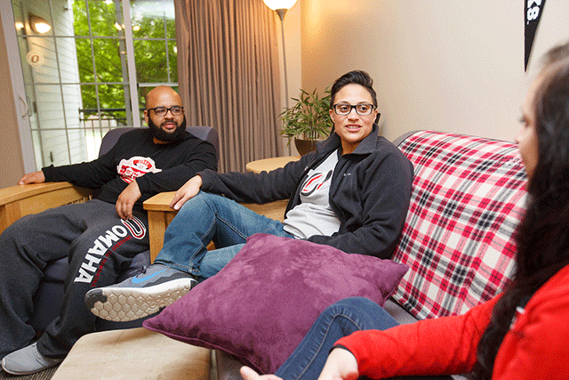 Three students sit and talk in a relaxed manner while seated on couches and chairs near a window.