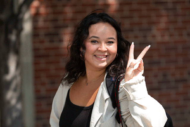 Smiling college student with a backpack stands in front of a campus building, looking at the camera and making a peace sign.