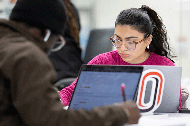 A student wearing glasses focuses on a laptop with a UNO logo sticker while studying at a table with others nearby.