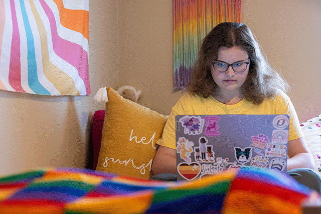 a female student wearing glasses sits on her bed in a colorful room looking at her laptop.