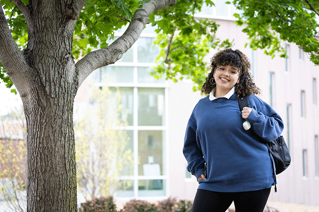 A smiling student with a backpack stands outdoors near a tree on campus, with a building in the background.