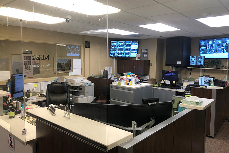 Reception area of a testing center with desks, computers, printers, and multiple wall-mounted monitors displaying camera feeds.