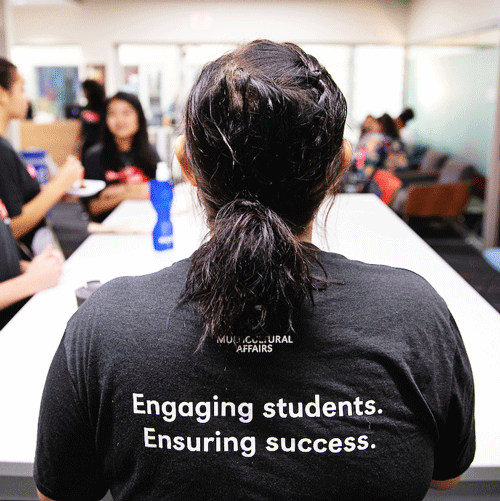 Multicultural Affairs staff member showing back of t-shirt that reads ensuring student success