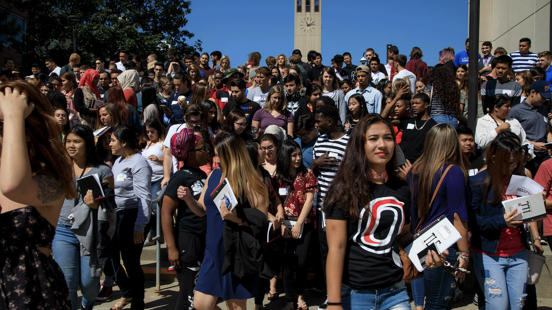 A large, diverse group of college students stand closely together on outdoor campus steps under a bright blue sky. Many are holding notebooks or folders and wearing name tags, suggesting an orientation or campus event. A clock tower rises in the background, and sunlight casts strong shadows across the crowd as students look in different directions and talk among themselves.