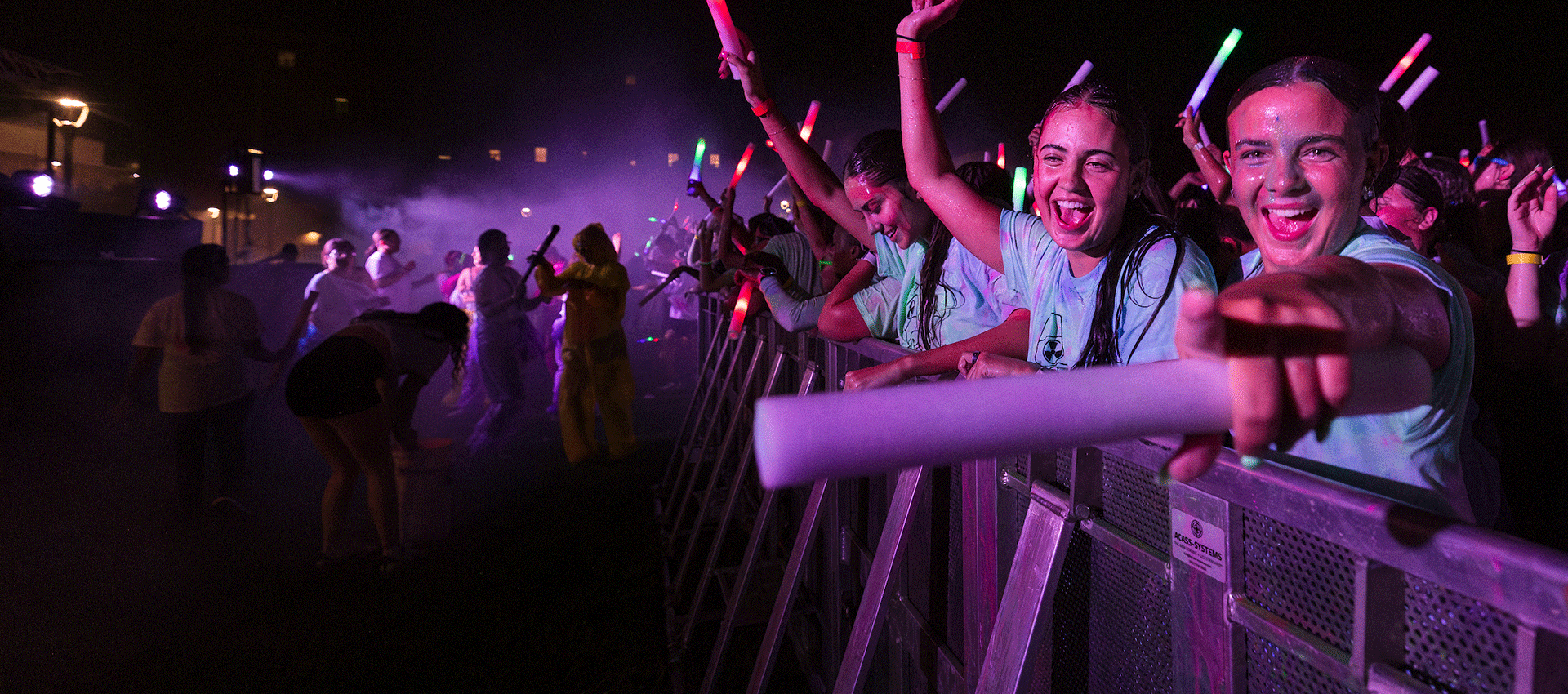 A crowd of excited young people at a nighttime outdoor event, splashed with colorful powder and holding glowing light sticks. Several smiling participants at the front rail reach toward the camera as bright stage lights and mist create a vibrant, energetic atmosphere.