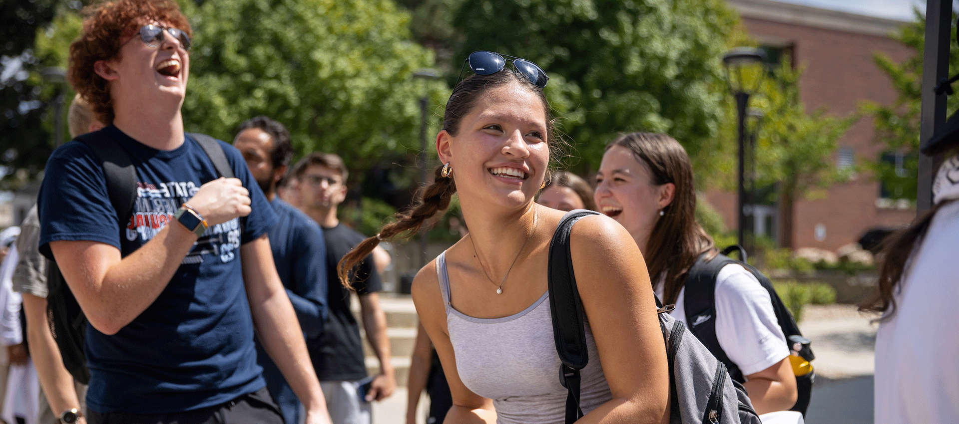 a female student in a tank top and wearing a backpack is smiling and looking at a group of people.