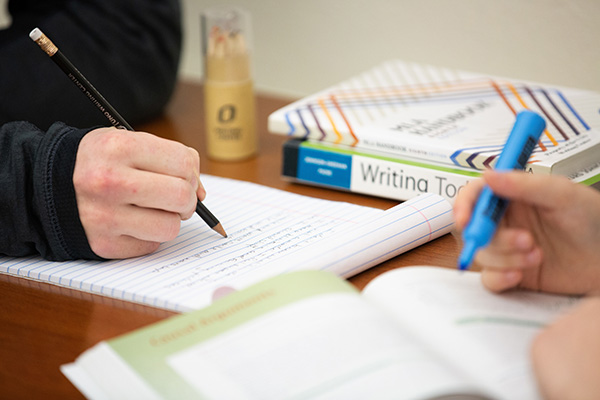 a hand writing in a notebook with writing books stacked to the side