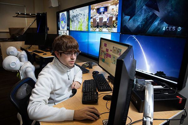 a student wearing glasses works on a computer with artificial intelligence robots behind him