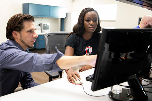 a man points to a computer screen as he teaches a young woman about computer science