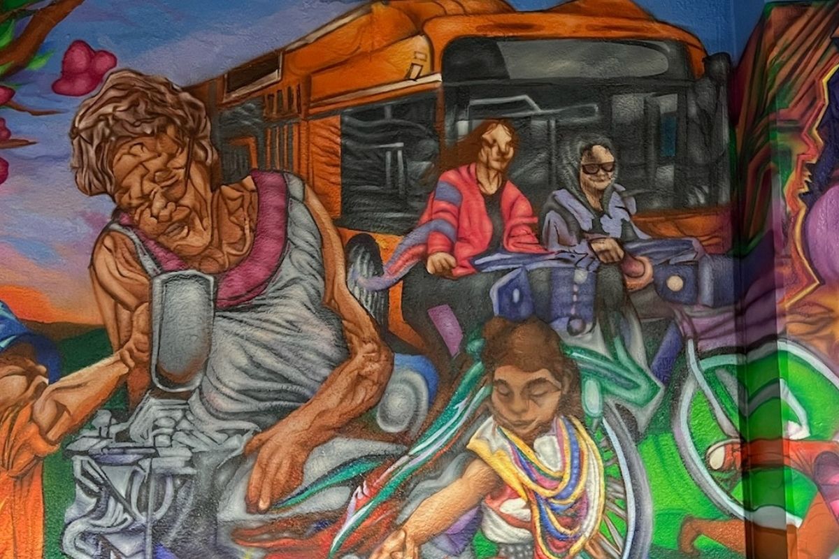 **Alt text:** Detail from *Community Visions II,* a mural by Hugo Zamorano (2025), showing vibrant figures of community members, including a seamstress, a child, and people riding a city bus, symbolizing connection, labor, and everyday life in Omaha.