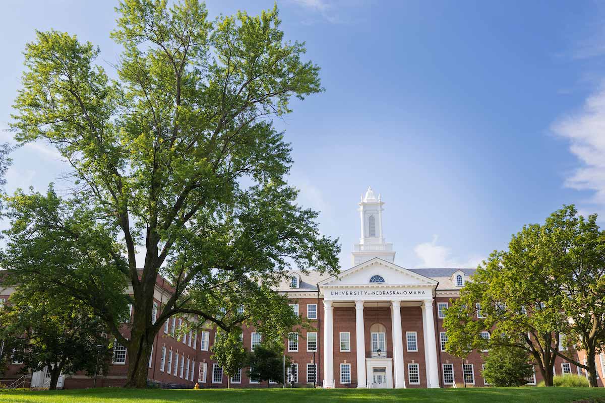 A red-brick academic building with tall white columns and a central cupola, framed by large leafy trees under a clear blue sky. 