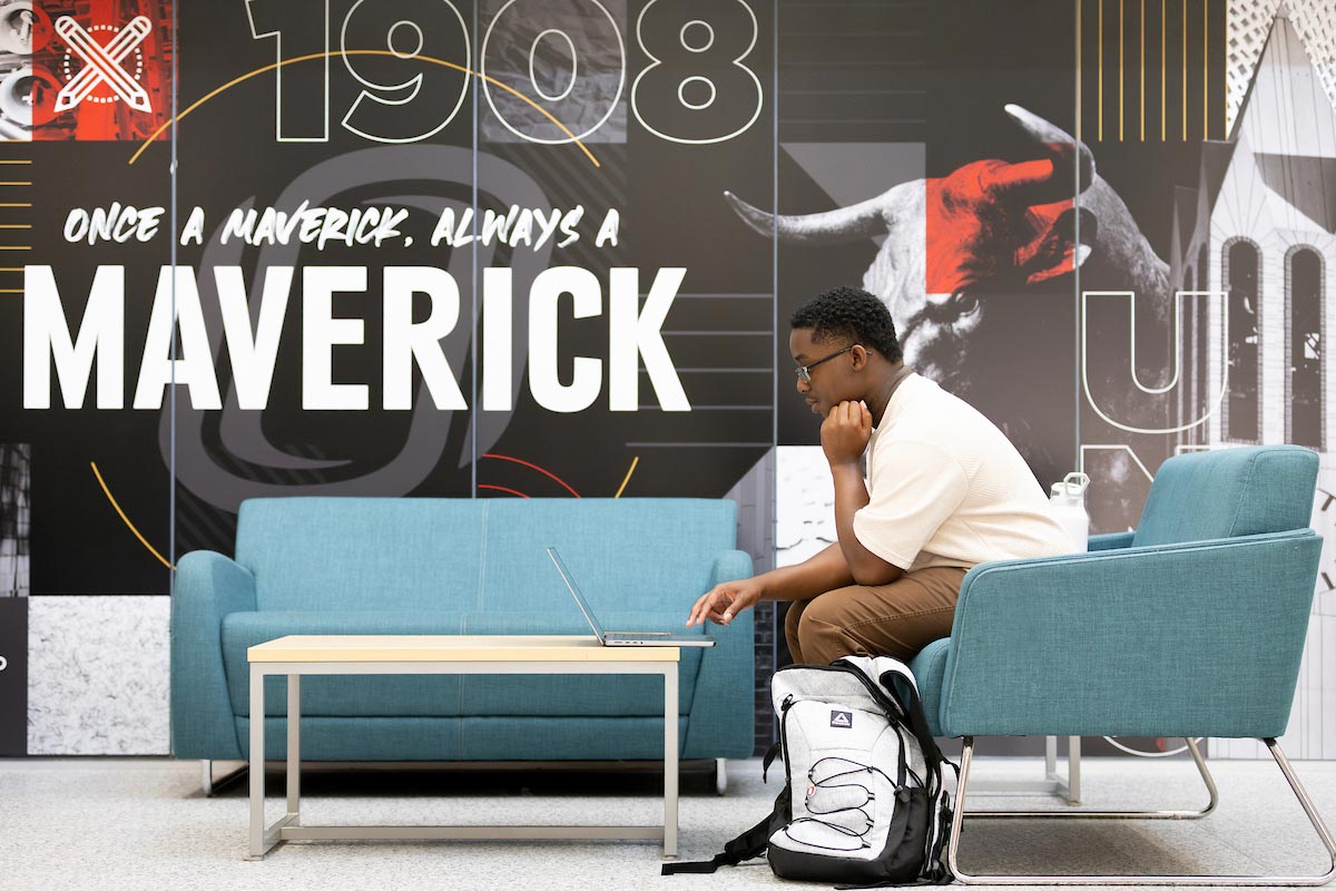 A University of Nebraska at Omaha student sits in a blue chair with a laptop, studying at a table in the Milo Bail Student Center. A large wall display behind him reads “Once a Maverick, Always a Maverick” with UNO graphics and the year 1908. 