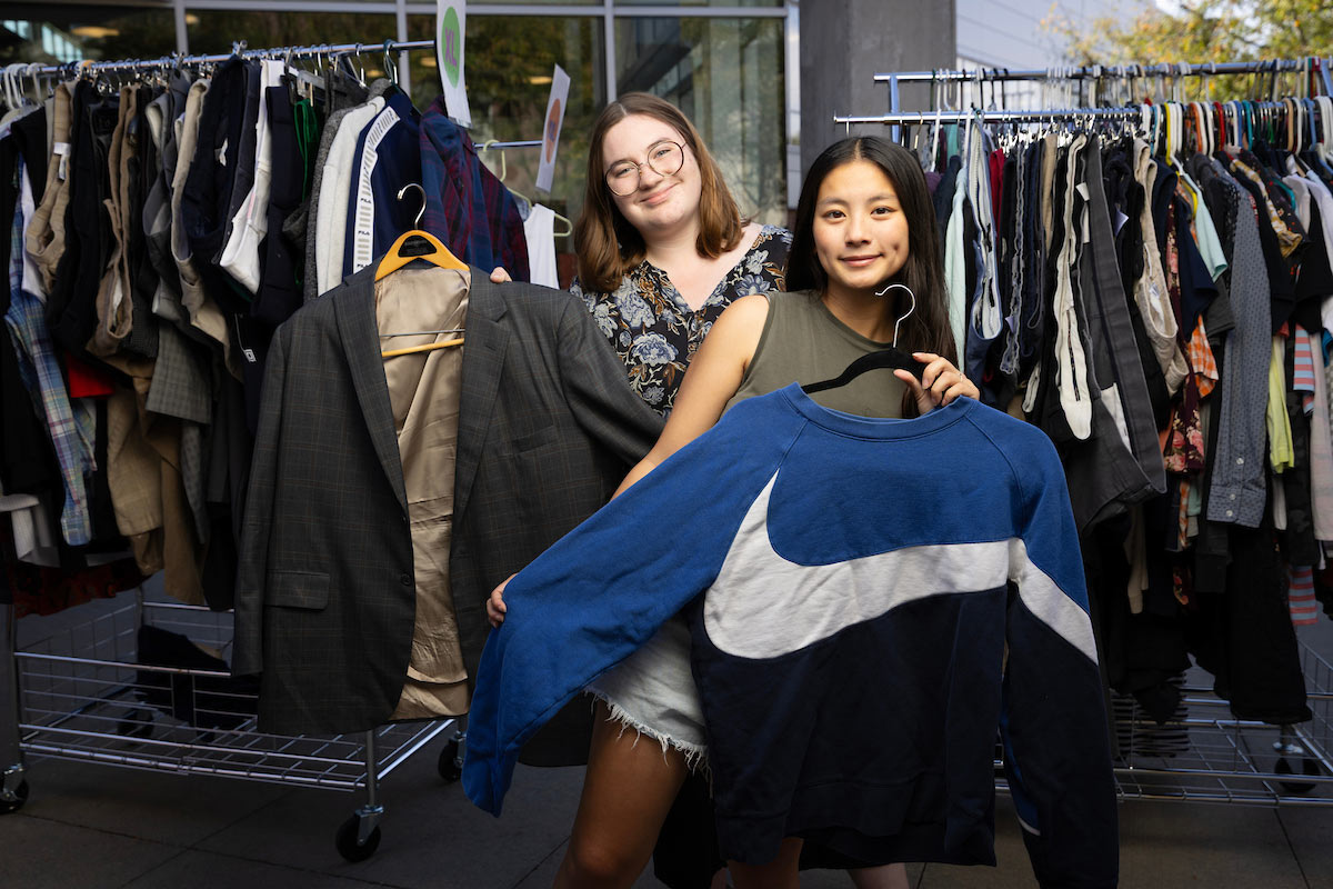 Two students hold up clothing on hangers in front of clothing racks at a Durango’s Outfitters pop-up, one with a suit jacket and the other with a blue Nike sweatshirt.